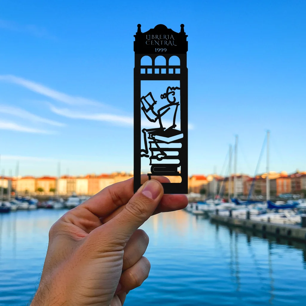 Mano sujetando el marcapáginas troquelado de la libreria La Central de Gijón con el puerto deportivo de fondo